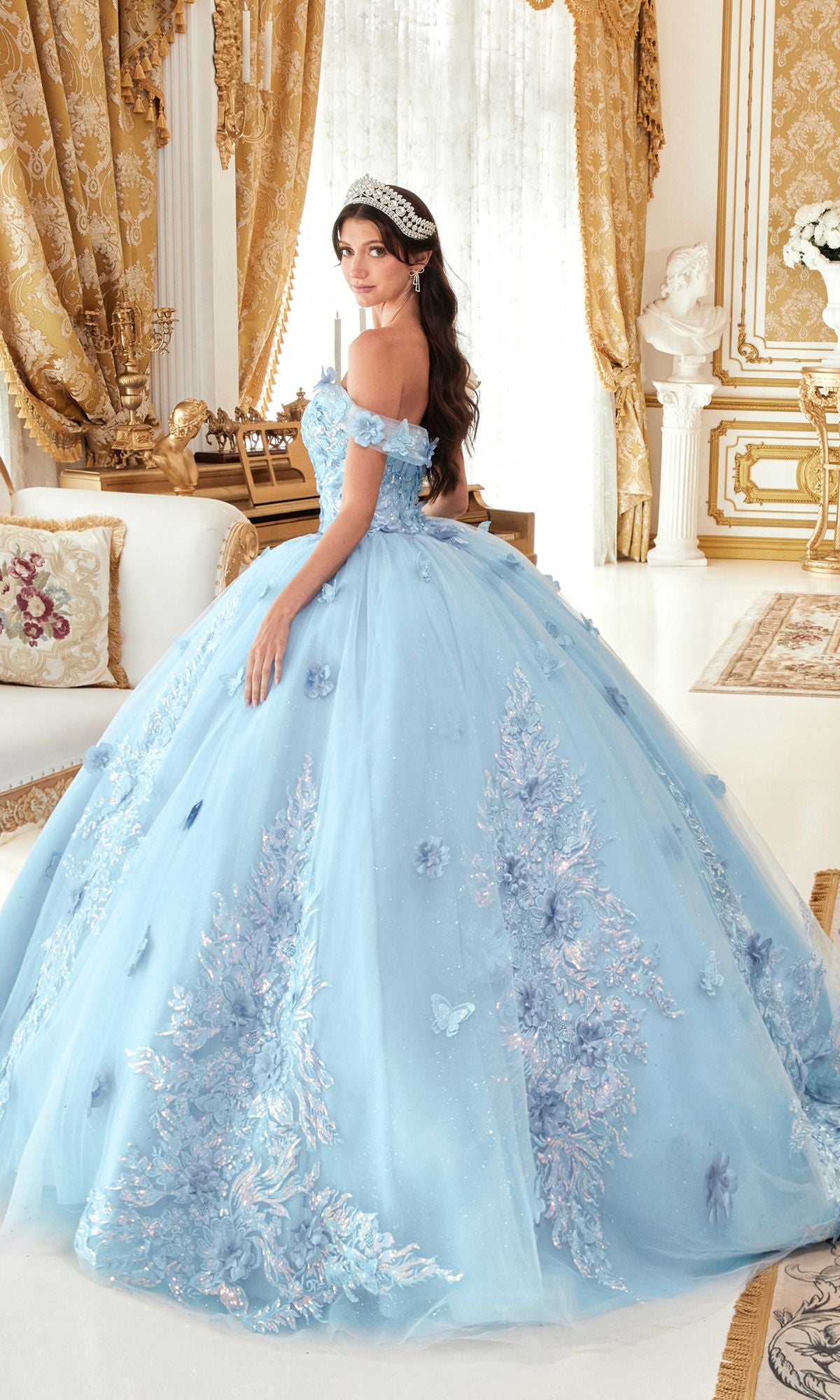 A woman wearing the Quinceanera Dress 15713 by Ladivine, featuring blue off-shoulder design with floral and butterfly appliques, stands in an ornate, gold-accented room, looking over her shoulder.